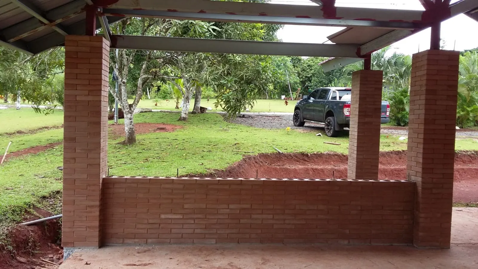 Muro bajo y tres columnas levantados con ladrillo ecológico color terracota, bajo un techo metálico y frente a un jardín